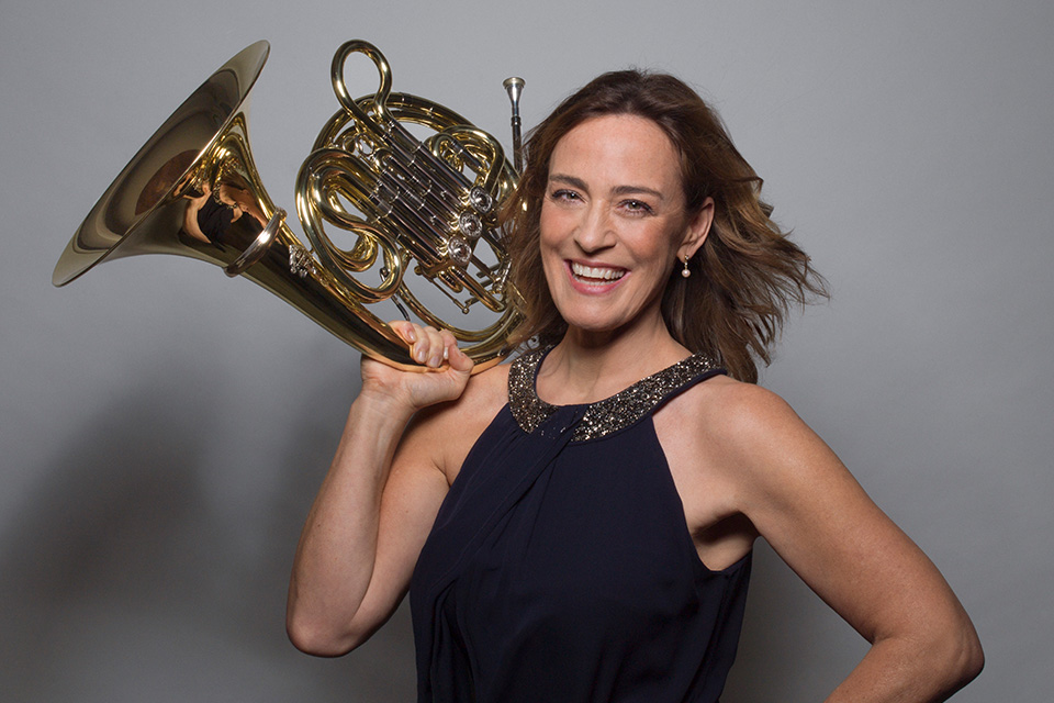 Sarah Willis holds her horn over her right shoulder with her other hand on her hip, smiling