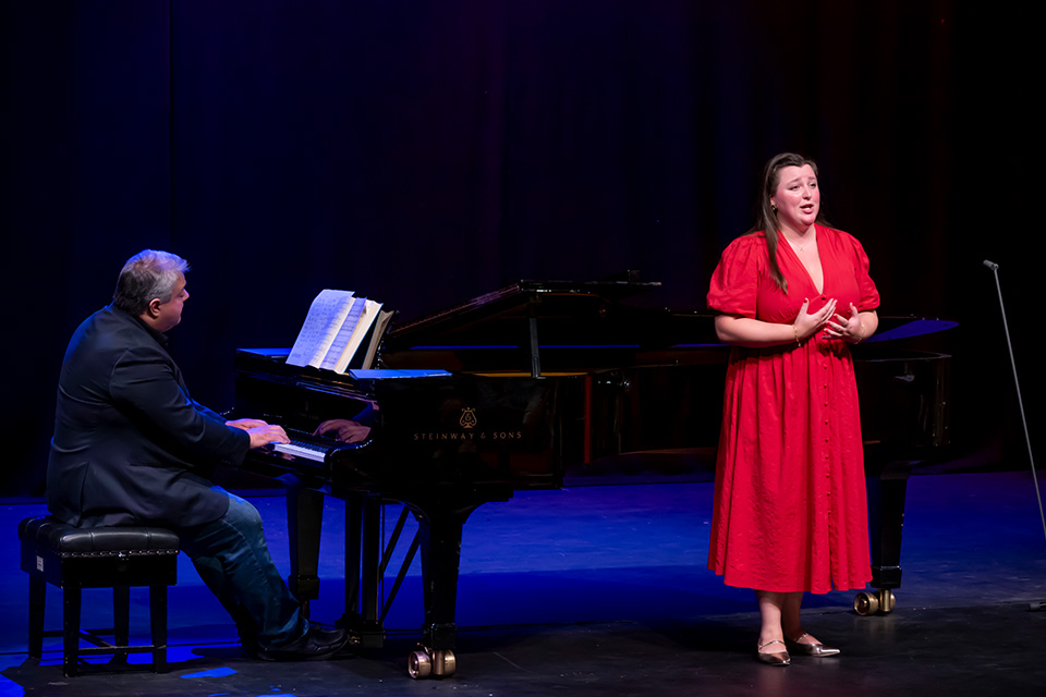 A singer wearing a red dress performs on stage, a pianist accompanying behind her