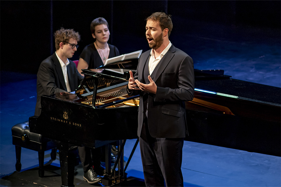 A vocalist sings emotionally, a pianist accompanying behind him on a brightly lit stage