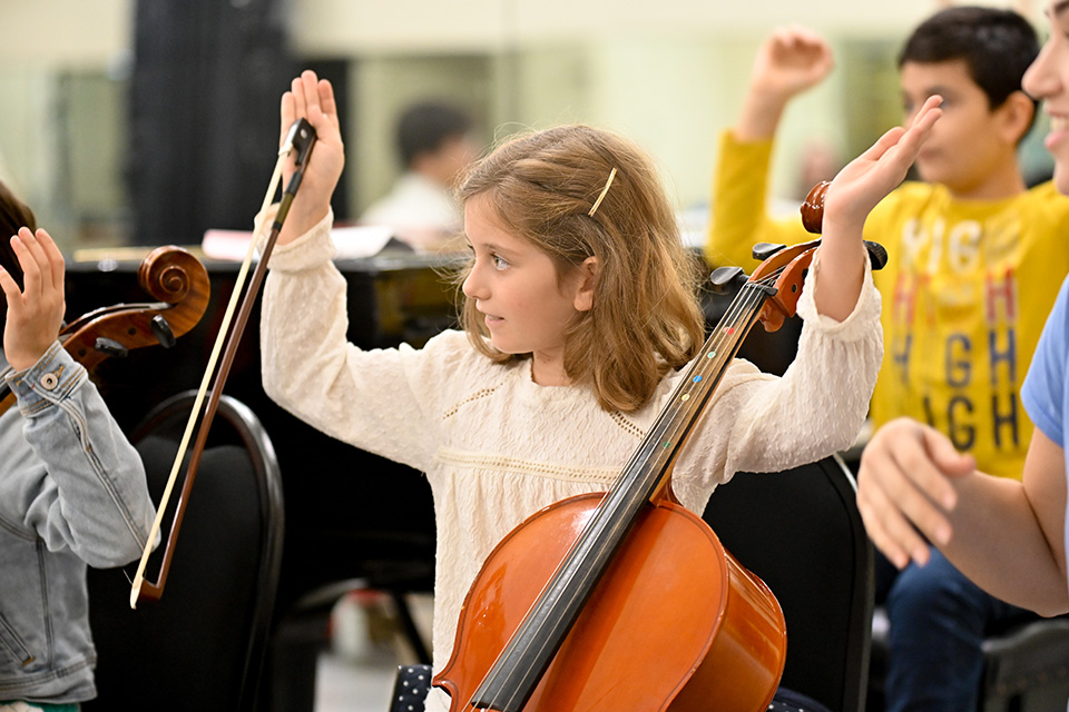 A young girl rests her cello on her knee with her hands in the air