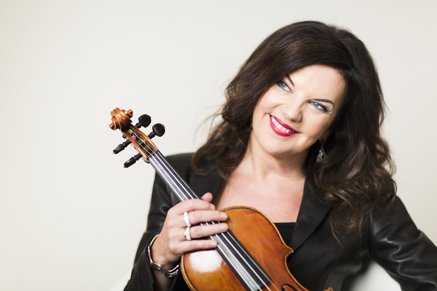 A photograph of Tasmin Little wearing black, holding her violin and smiling, looking off to the side