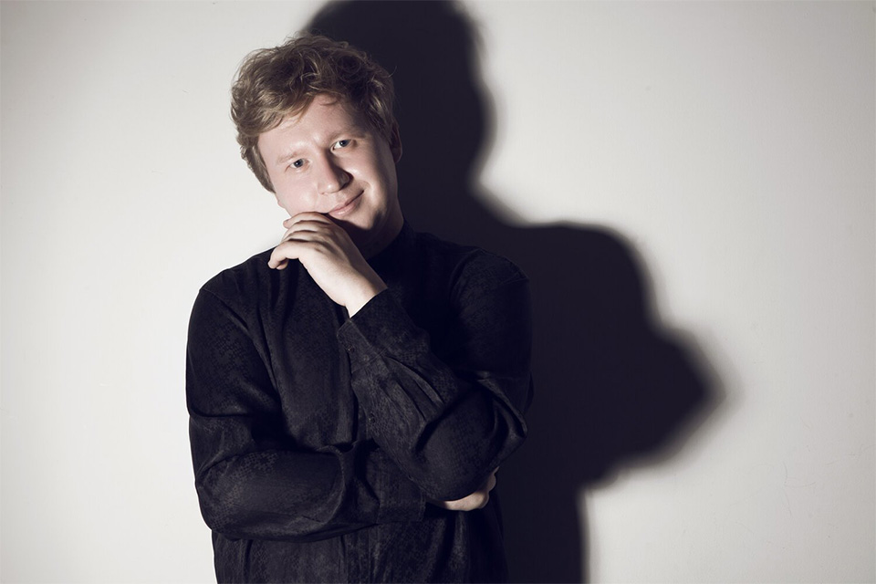 Yury Shadrin wears a black shirt rests his chin on his hand, his shadow large on the white wall behind him