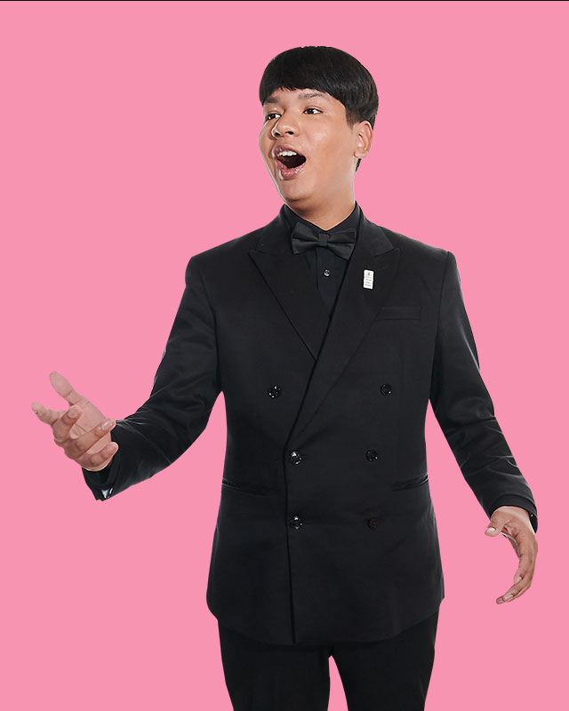 A man wearing a black double-sided blazer, with short hair, singing, with a light pink background.