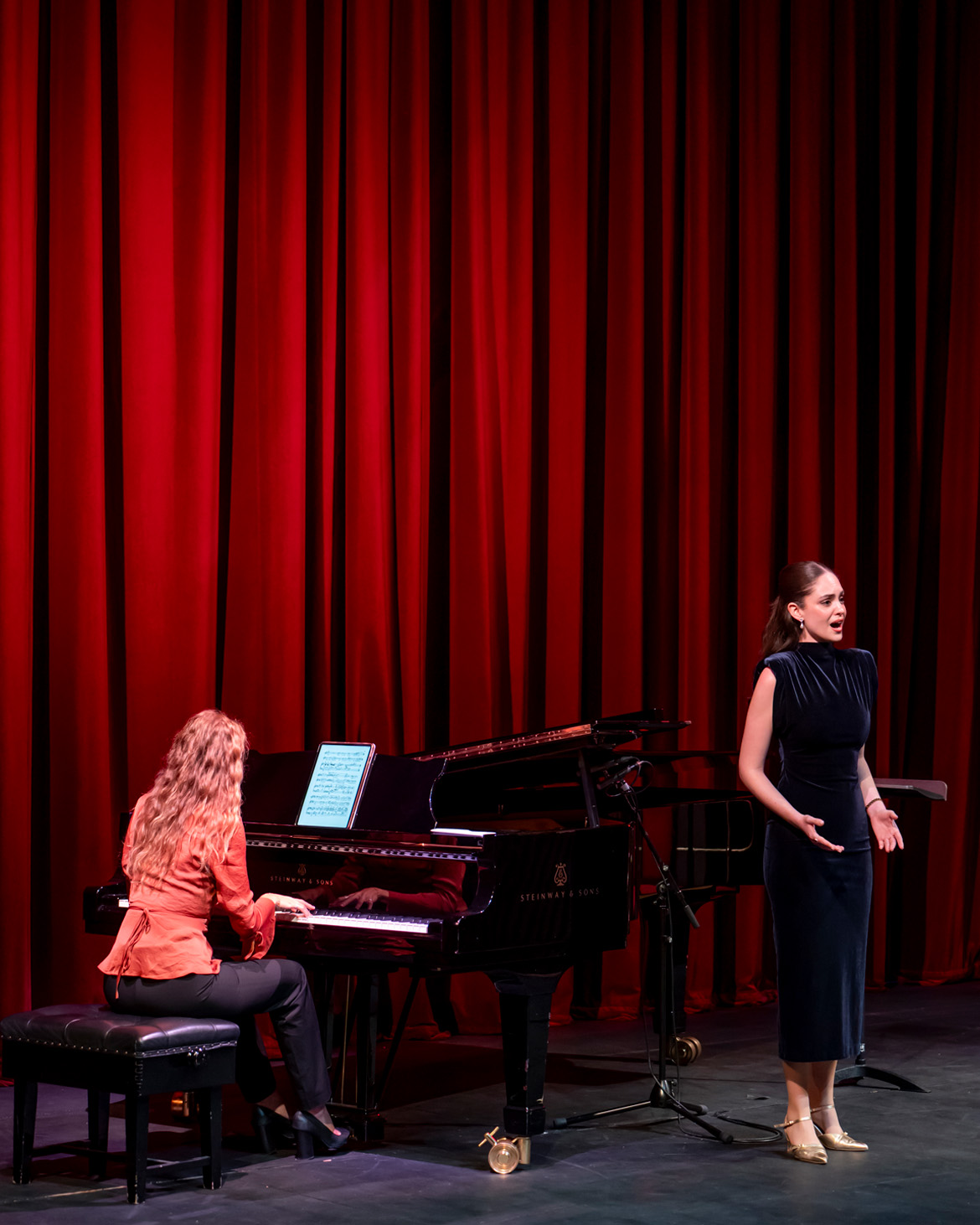 A singer and piano accompanist, performing on stage, with a red curtain behind.