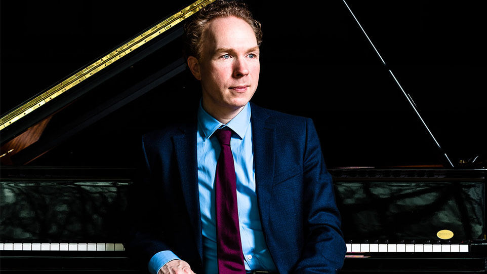 Ralph van Raat: A man dressed in a suit and tie sits next to a piano, suggesting a formal or musical setting.