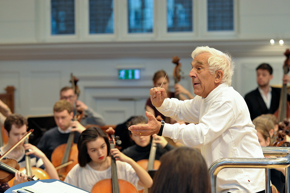 Vladimir Ashkenazy holding a baton and conducting in a concert in the Amaryllis Fleming Concert Hall with cellists in view