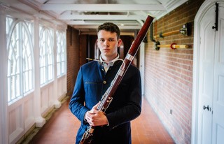 a man with a bassoon, wearing a navy jacket and standing against a corridor background